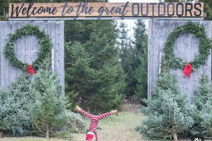 Child in striped outfit doing cartwheel under sign 'Welcome to the great OUTDOORS' with wreaths and pine trees.