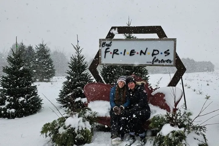 Two people sit on a snowy bench with a sign reading 'Best FRIENDS forever,' surrounded by snow-covered trees.