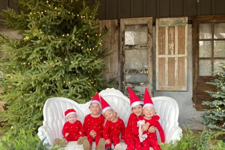 Five children in red elf outfits sit on a white sofa by Christmas trees and rustic doors.