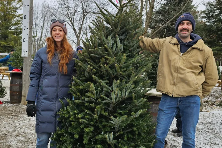 Two people posing with a Christmas tree outdoors on a snowy day.