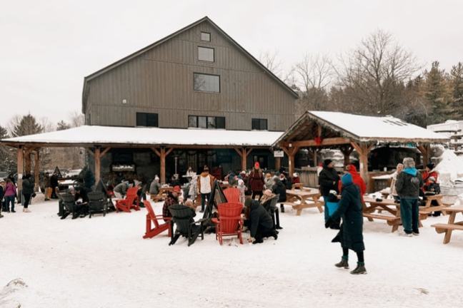 People gather outside a barn in winter, with snow-covered ground and seating.