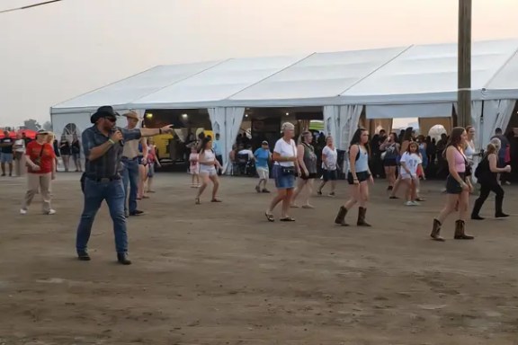 People line dancing in an outdoor area with a large tent in the background.