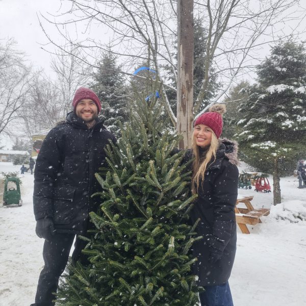 Two people in winter clothes stand beside a Christmas tree in a snowy setting.