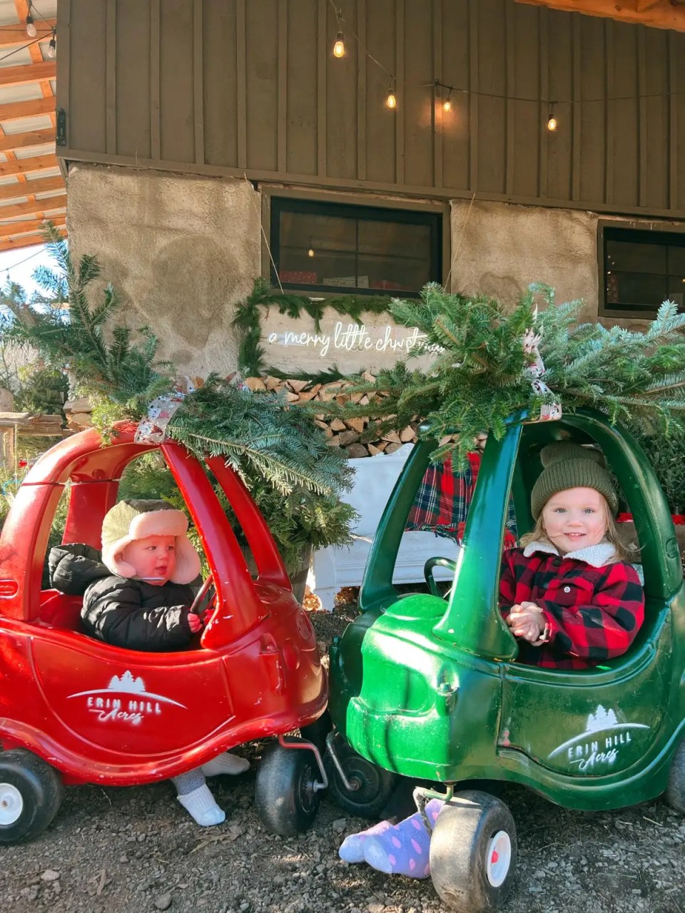 Two children in red and green toy cars decorated with pine branches.