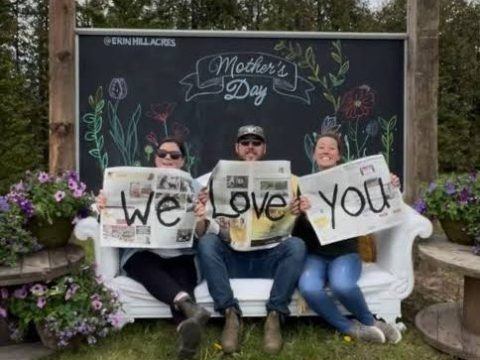 Three people on a bench holding 'We Love You' signs by a Mother's Day chalkboard.